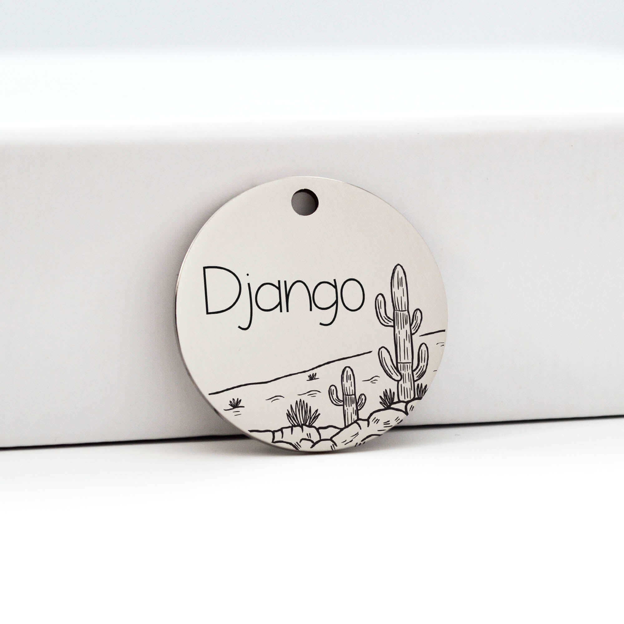 Round silver pet ID tag with the name 'Django' engraved in black, featuring a desert landscape design with cactus illustrations, resting against a white box.