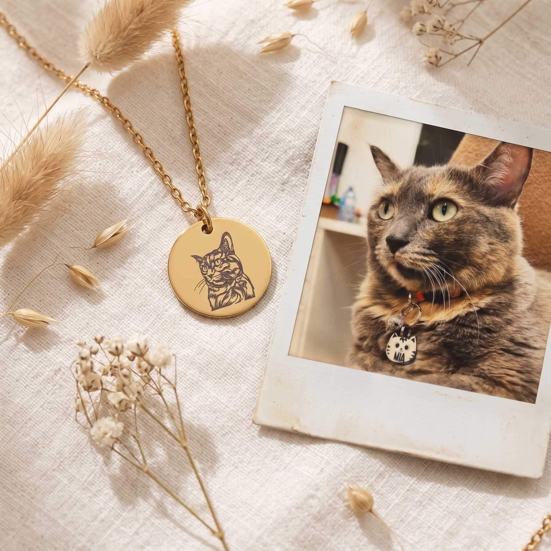 A gold round pendant necklace engraved with a cat profile, displayed on a textured cream linen fabric alongside dried pampas grass, baby's breath, and a Polaroid-style photo of a tortoiseshell cat wearing a collar.