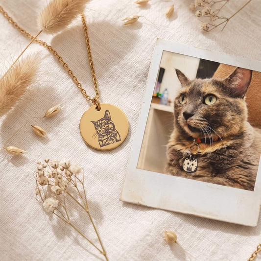 A gold round pendant necklace engraved with a cat profile, displayed on a textured cream linen fabric alongside dried pampas grass, baby's breath, and a Polaroid-style photo of a tortoiseshell cat wearing a collar.