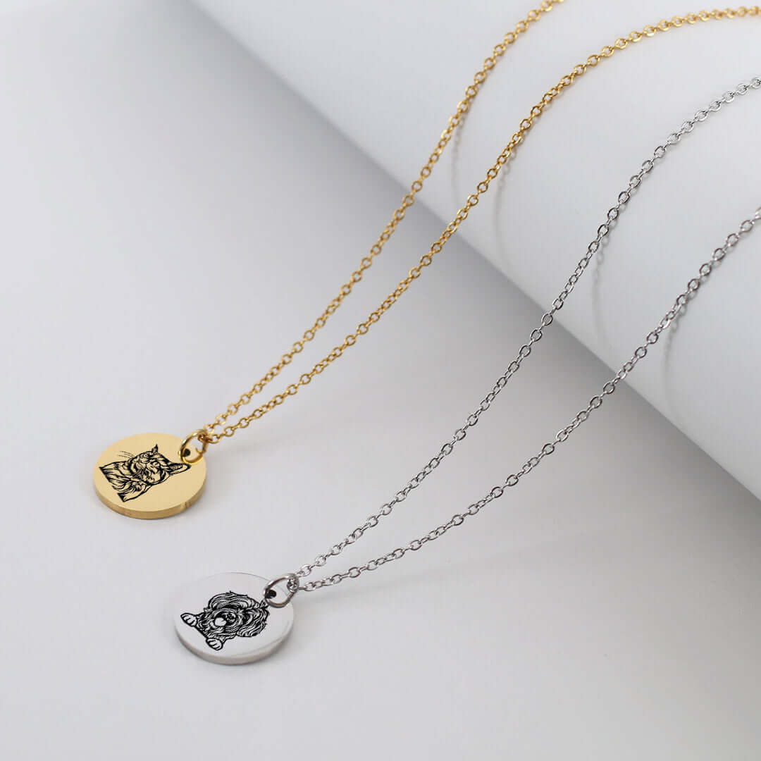 Two round pendant necklaces on delicate chains, one gold-toned and one silver-toned, each featuring an engraved cat face design, displayed on a white cylindrical prop against a light gray background.