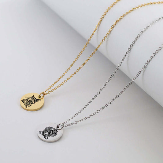Two round pendant necklaces on delicate chains, one gold-toned and one silver-toned, each featuring an engraved cat face design, displayed on a white cylindrical prop against a light gray background.