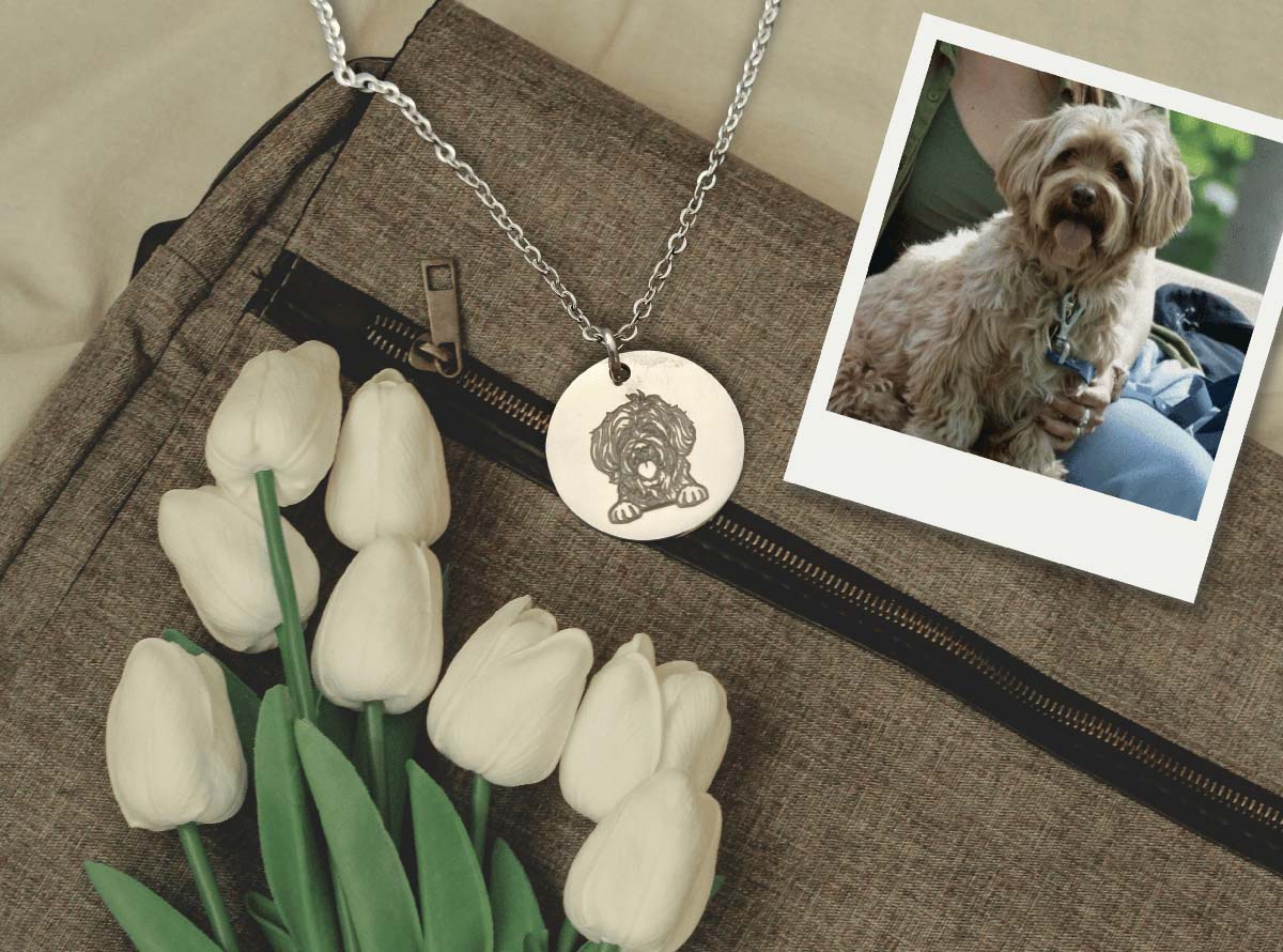 A silver round pendant necklace with an engraved dog design, displayed on a brown textured bag next to white tulips, with a small inset photo of a dog wearing a similar necklace.