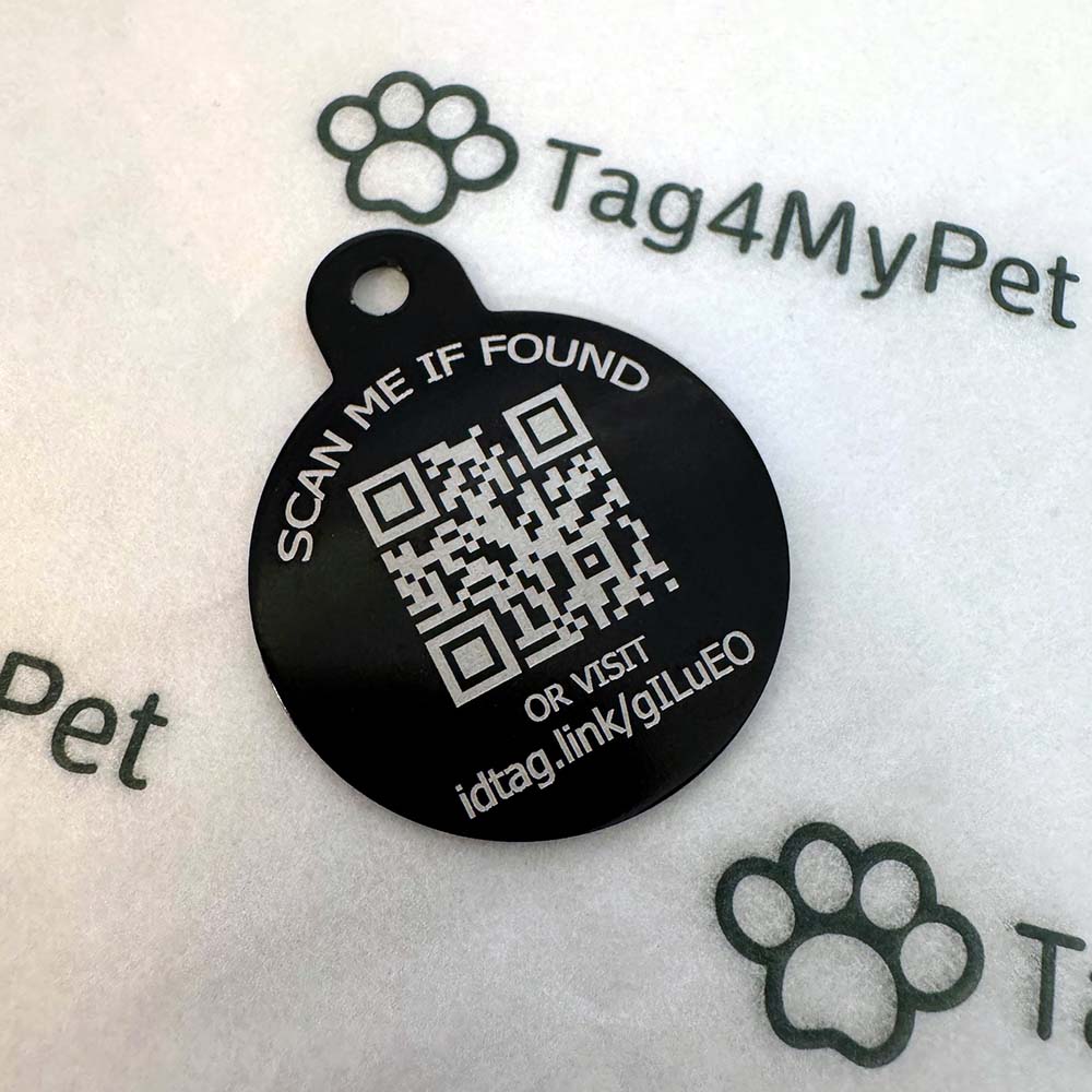 A round black pet ID tag with a white QR code and text reading 'SCAN ME IF FOUND' and 'OR VISIT idtag.link/gluEO', placed on a white surface with faint Tag4MyPet branding.