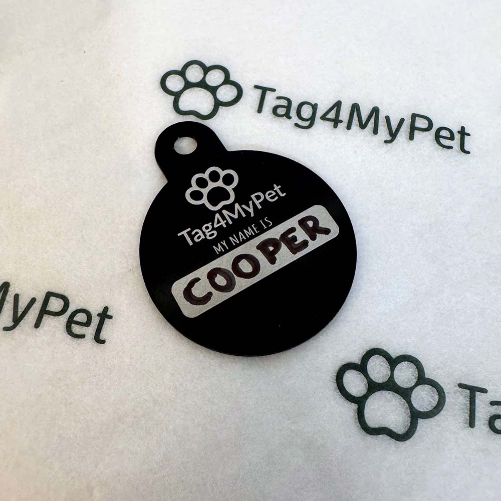 A round black pet ID tag with a paw print logo and the text 'Tag4MyPet', 'MY NAME IS', and 'COOPER' in white and brown lettering, placed on a white textured surface with faint watermark graphics.